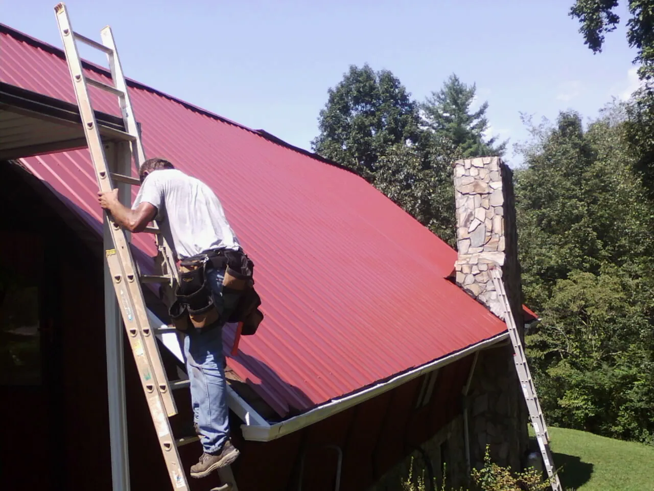 Roofer on a ladder beside a red metal roof and stone chimney — Soffit & Fascia Repair by Roofing Brothers in Oakwood